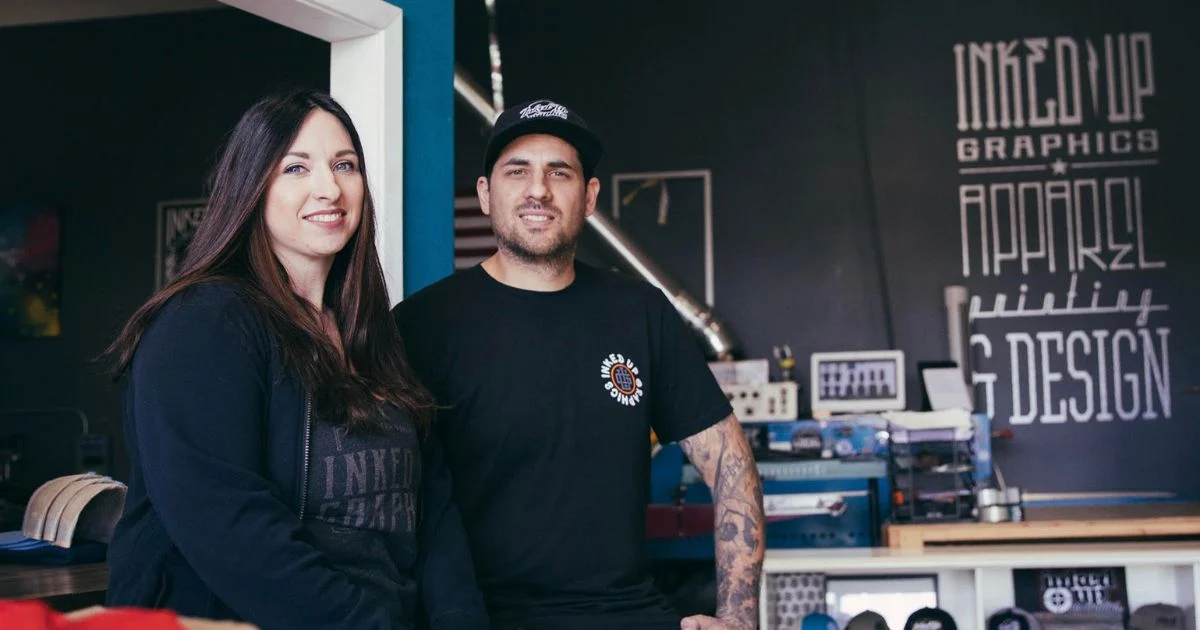 Inked Up Graphics' founders in their shop, the Inked Up Graphics apparel printing and design sign painted on the wall behind them