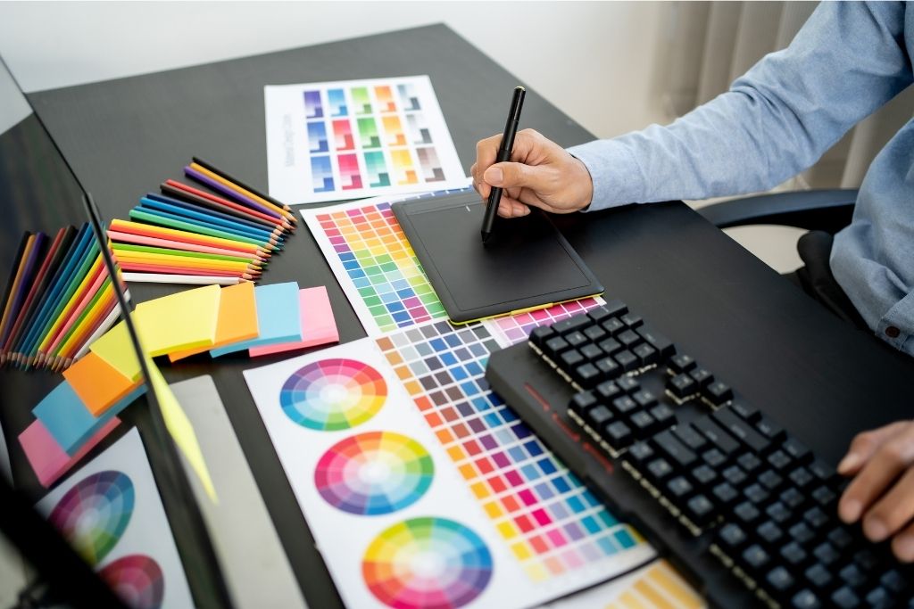 Person drawing on a drawing tablet with one hand while the other hand is on a keyboard, surrounded by color swatches, color pencils, and post-it notes.