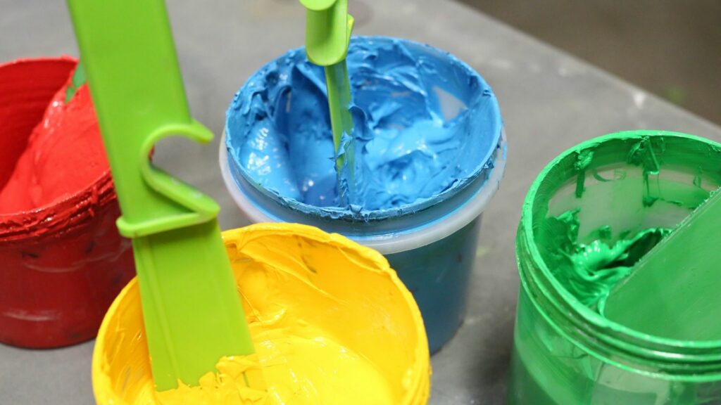 A pot of red ink, a pot of blue ink, a pot of yellow ink, and a pot of green ink are next to each other on a grey surface.