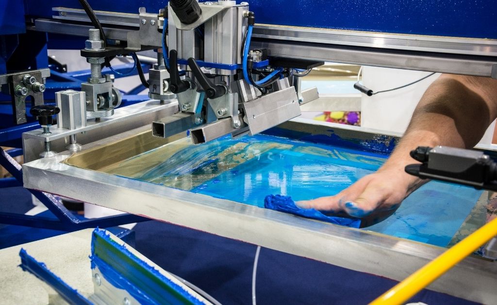 Person wiping blue ink off of an automatic screen printing press.
