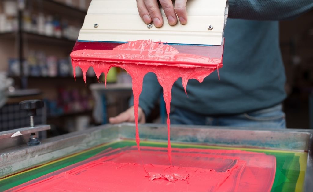 A person holding a squeegee that's dripping red ink onto a screen.